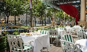 Utomhusservering med vita bord och stolar samt grönrandiga dynor, omgiven av grönska och ett glasräcke. (AI-genererad text)