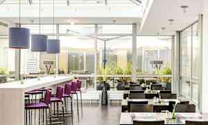 En restaurang med ljusa inredningsdetaljer, lila barstolar och mörka bord. Glasväggar släpper in naturligt ljus. (AI-genererad text)