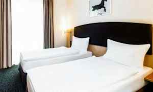 Två enkelsängar står bredvid varandra i ett hotellrum. Väggarna är ljusa och det finns ett konstverk av en häst ovanför huvudgaveln. (AI-genererad text)