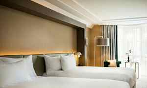 Ett hotellrum med två enkelsängar, en grön fåtölj, ett sidobord med dekor och en golvlampa, bredvid ett fönster med ljusa gardiner. (AI-genererad text)