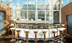 En cocktailbar med höga stolar framför en glasvägg med utsikt över en byggnad med färgglada takdekorationer. (AI-genererad text)