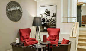 Två röda fåtöljer står runt ett runt bord, bredvid en golvlampa och framför en vägg prydd med konst i ett hotellinteriör. (AI-genererad text)
