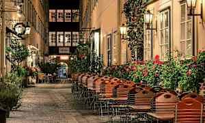 En pittoresk gränd med stolar och bord utanför restauranger, omgivna av blommande buskar och lampor. (AI-genererad text)