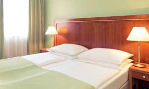 Ett hotellrum med en dubbelsäng som har vita sängkläder och gröna filtar. Två sänglampor står på träbord bredvid sängen. (AI-genererad text)