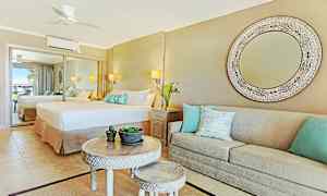 Ett elegant hotellrum med en stor spegel, en bred säng och en beige soffa med dekorativa kuddar. Ett runt bord med mindre bord och växtdekorationer i mitten. (AI-genererad text)