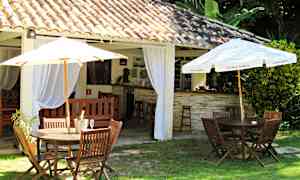 Utomhusmiljö med bord och stolar av trä samt parasoller, intill en byggnad med tegelpannor och invändiga gardiner. (AI-genererad text)