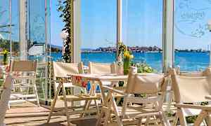 Utsikt från en restaurang med vita stolar och bord, med glasväggar som erbjuder vyer över havet och en kuststad. (AI-genererad text)