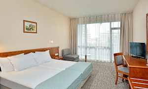 Ett hotellrum med en dubbelsäng, skrivbord, stol, TV och sittgrupp. Ett stort fönster släpper in dagsljus. (AI-genererad text)