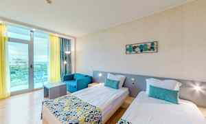 Ett hotellrum med två enkelsängar, en blå fåtölj, och ett bord. Rummet har ljusa väggar och ett stort fönster med gul gardin och balkong. (AI-genererad text)