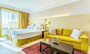 Ett hotellrum med en stor dubbelsäng, gul soffa, ett bord med dekorativ vas och en röd matta. (AI-genererad text)