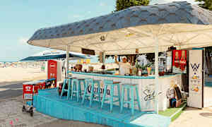 En strandbar med blåa stolar och tak, belägen nära sandstranden. (AI-genererad text)
