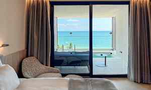 Ett hotellrum med en säng och fåtölj, samt utsikt över en balkong med havet i bakgrunden. (AI-genererad text)