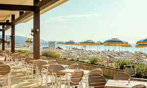 En uteservering med bord och stolar framför en strand med solstolar och parasoller. (AI-genererad text)