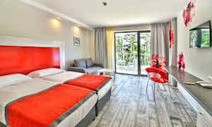 Ett hotellrum med två enkelsängar, en soffa, en skrivbordsplats och en balkong med utsikt mot grönska. (AI-genererad text)