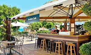 En kafé med bord och stolar utomhus vid Sunshine Café, med grönska och havsutsikt i bakgrunden. (AI-genererad text)