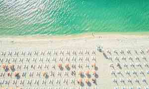 Flygfoto av en strand med parasoller och solstolar. Havet är färgstarkt grönblått. (AI-genererad text)