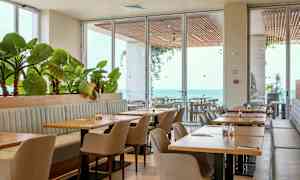 En restaurang med bord, stolar och växter vid stora fönster med utsikt över havet. (AI-genererad text)