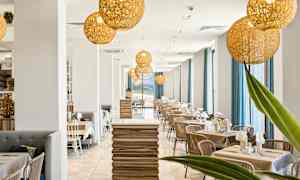 En modern restaurang med en ljus inredning innehållande träbord, stolar och stora fönster med blåa gardiner. Dekorativa belysningsarmaturer hänger från taket. (AI-genererad text)