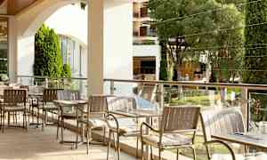 En utomhusservering med bord och stolar, omgiven av gröna träd och planteringar. (AI-genererad text)