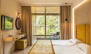 Ett hotellrum med en dubbel bädd, gul sänggavel, litet skrivbord, och en stor fönsterfront mot naturen. (AI-genererad text)