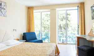 Rum med säng, blå fåtölj och skrivbord, med stora fönster mot balkong. (AI-genererad text)