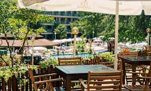 En uteplats med bord och stolar i trä under ett parasoll, vid en trädgård och poolområde. (AI-genererad text)