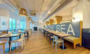 En modern restaurang med ljus inredning, träbord och stolar, samt texten 'LOVE THE SEA' på ett blått ryggstöd. (AI-genererad text)