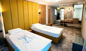 Ett hotellrum med två enkelsängar med gul klädsel och vita sängkläder. Rummet har en modern inredning med en gul väggpanel, ett skrivbord, sittmöbler och en liten förvaringshylla. (AI-genererad text)