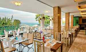 En restaurang med bord och stolar i ljusa färger, utsikt mot havet och omgiven av träd. (AI-genererad text)