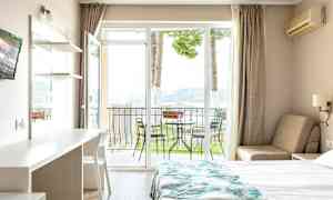 Ett hotellrum med en säng, ett bord och en stol, samt en balkong med utemöbler och utsikt mot naturen. (AI-genererad text)