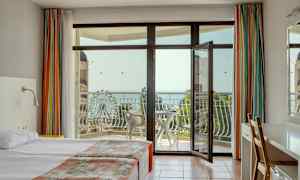 Hotellrum med dubbelsäng, öppen balkongdörr, och utsikt över havet och ett pariserhjul. (AI-genererad text)