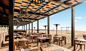 Utomhusrestaurang med träbord och stolar, belägen vid en sandstrand. (AI-genererad text)