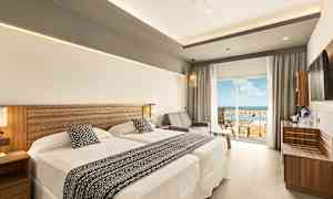 Ett hotellrum med två sängar och en sittgrupp bredvid en glasdörr som leder till en balkong med utsikt. (AI-genererad text)