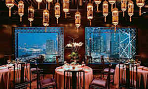 Ett elegant inrett restaurangrum med runda bord, stolar och lyktor i taket, med utsikt över en stadsmiljö genom fönstren. (AI-genererad text)