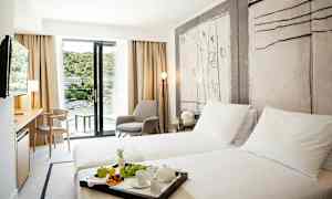 Ett hotellrum med två enkelsängar, en sittgrupp och en balkong med utsikt över en grönskande omgivning. (AI-genererad text)