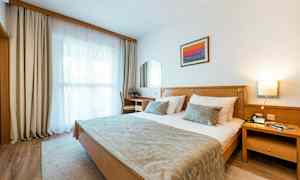 Ett hotellrum med en dubbelsäng, en skrivbordsstol, ett fönster med gardiner och en väggprydnad. (AI-genererad text)