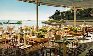 Uteservering vid havet med bord och stolar under ett tak, omgiven av växter och en strand i bakgrunden. (AI-genererad text)