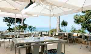 En uteservering med vita parasoller, bord och stolar samt utsikt över havet. (AI-genererad text)