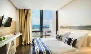 Ett hotellrum med en dubbelsäng, sängkläder i blå- och vitrandigt mönster, och en stor glasvägg som visar en balkong med utsikt över havet. Rummet har även en TV och skrivbord. (AI-genererad text)