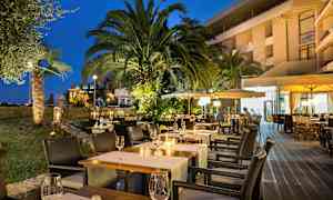 Kvällsbild av en utomhusrestaurang med upplysta bord, omgiven av palmer och grönska. (AI-genererad text)