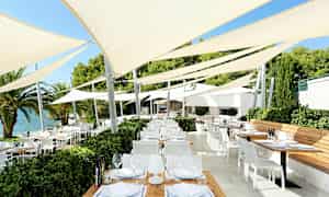 Ett utomhusområde med flera bord och stolar under spännade parasoll nära vattnet. (AI-genererad text)