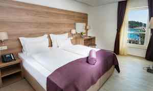 Ett hotellrum med en dubbelsäng, vita lakan och lila överkast. Det finns ett skrivbord med en stol och en spegel. Fönstret visar utsikt mot havet. (AI-genererad text)