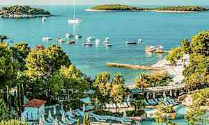 Utsikt över en poolanläggning med solstolar omgiven av grönska. I bakgrunden syns en strand med båtar och en vik med små öar. (AI-genererad text)