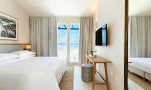 Ett hotellrum med en stor säng, ett skrivbord, och en TV på väggen. Stora fönster med utsikt mot havet. (AI-genererad text)