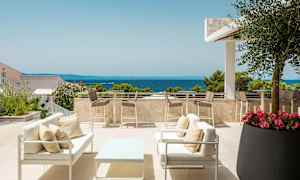 En terrass med sittmöbler och bord samt utsikt över havet. Det är omgiven av växter och träd med klar himmel. (AI-genererad text)