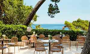 Bild av en utomhusservering med bord och stolar, omgiven av grönska och med havsutsikt. (AI-genererad text)