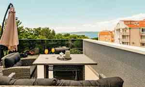 Terrass med bord och stolar, orange drycker i glas, paraply och utsikt över träd, byggnader och hav. (AI-genererad text)