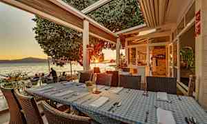 Restaurangbord med blåvitrutiga dukar på en terrass vid havet under solnedgången. (AI-genererad text)