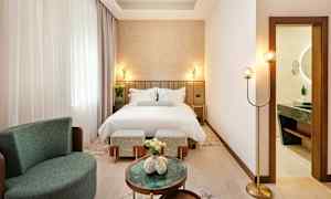 Ett elegant hotellrum med en bäddad säng, fåtölj och små bord. Ett badrum syns genom en öppnad dörr. (AI-genererad text)