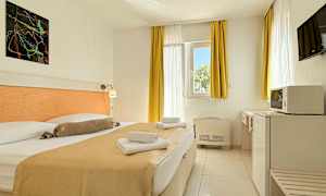 Ett hotellrum med en dubbelsäng, ljusa gardiner och möbler samt ett fönster som släpper in dagsljus. (AI-genererad text)